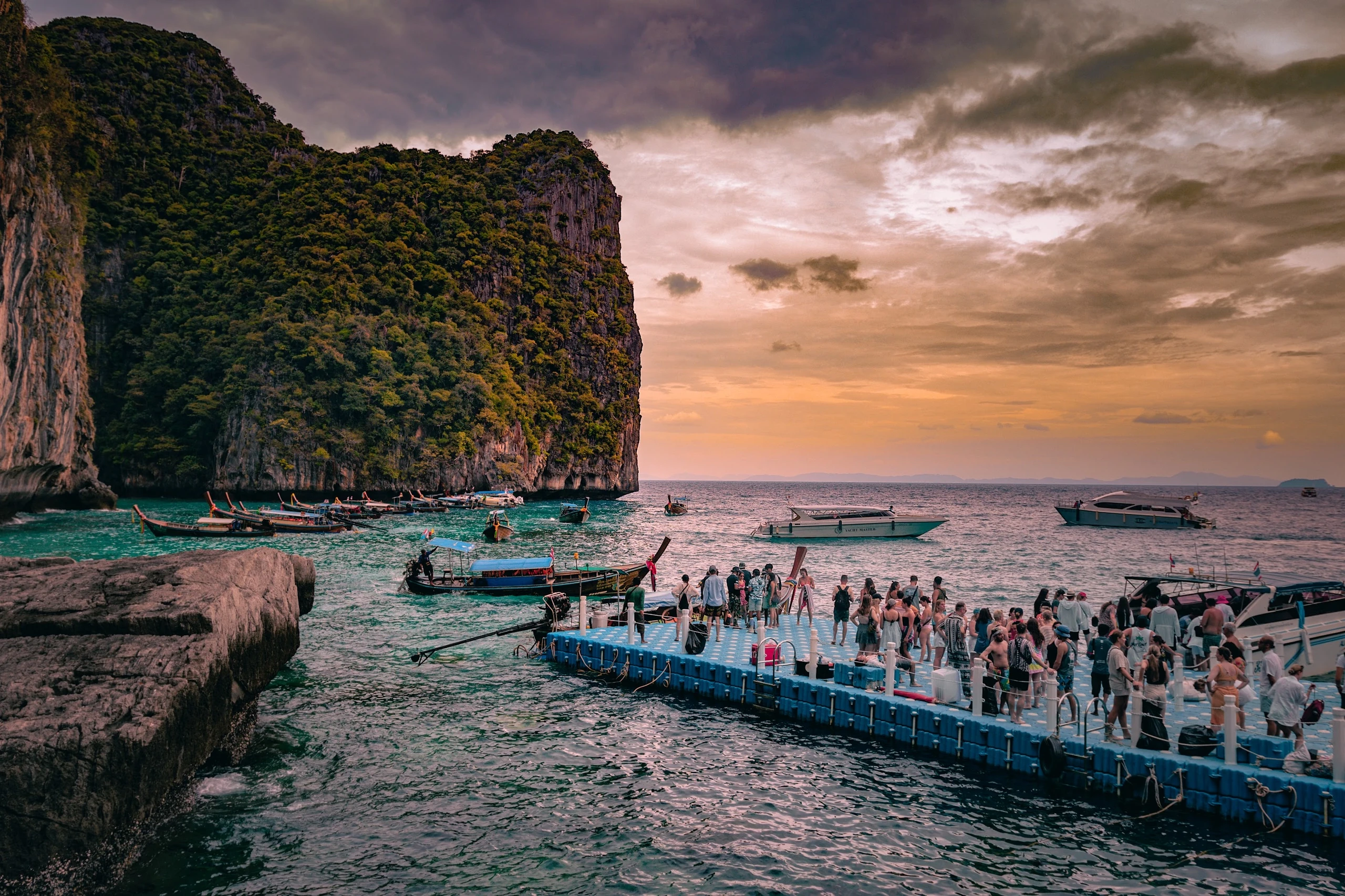 Long-tail boats gathered beside a limestone cliff beneath a pink sunset sky.