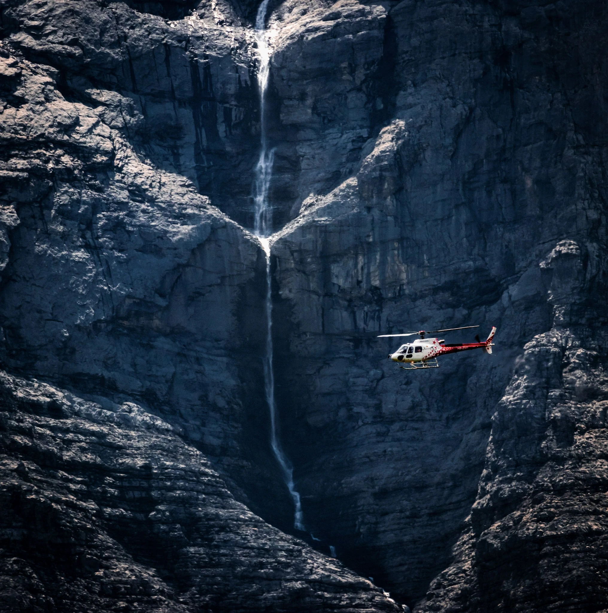 A helicopter passing a high waterfall on a dark, sheer mountain face.