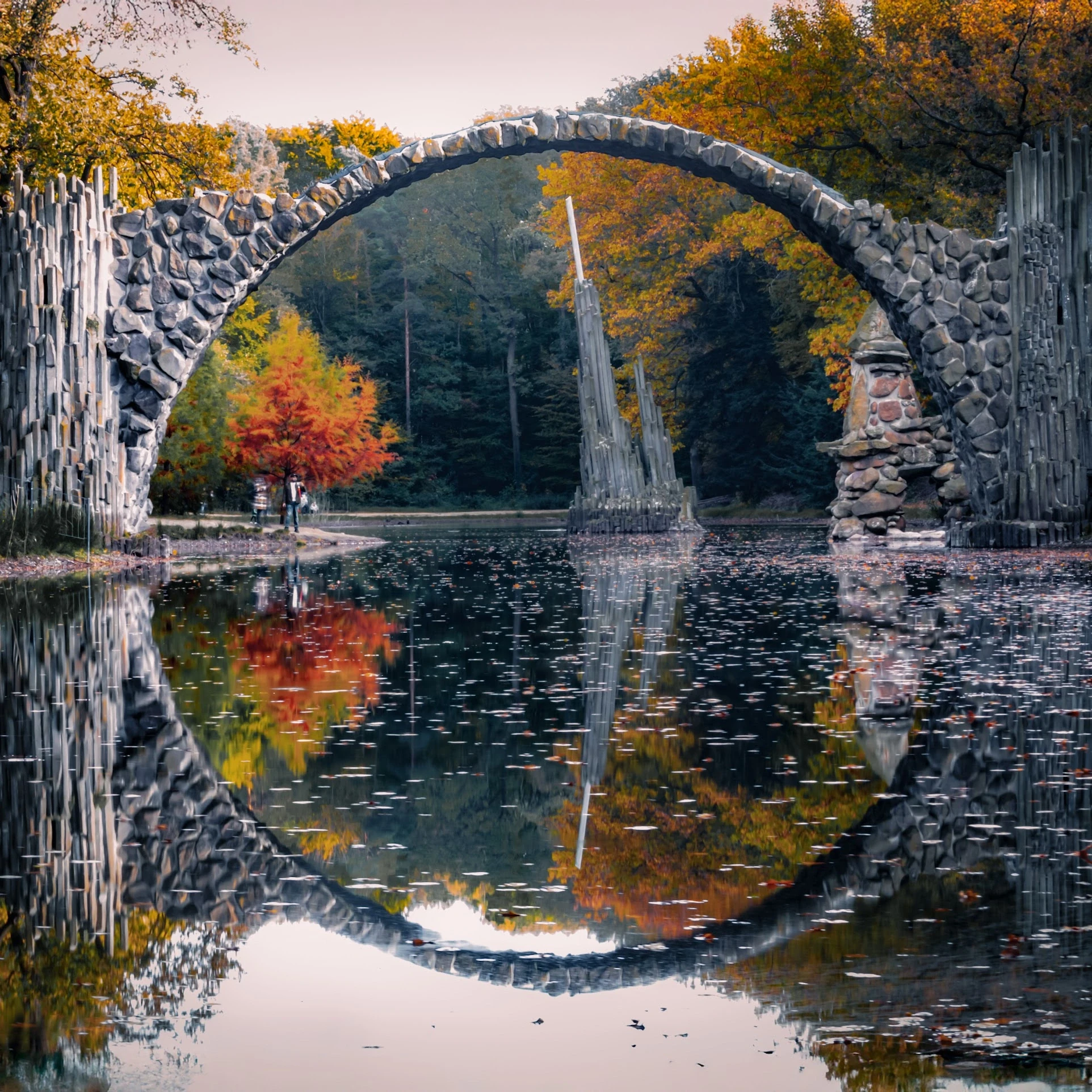 A stone arch bridge forming a circle with its reflection among autumn trees.