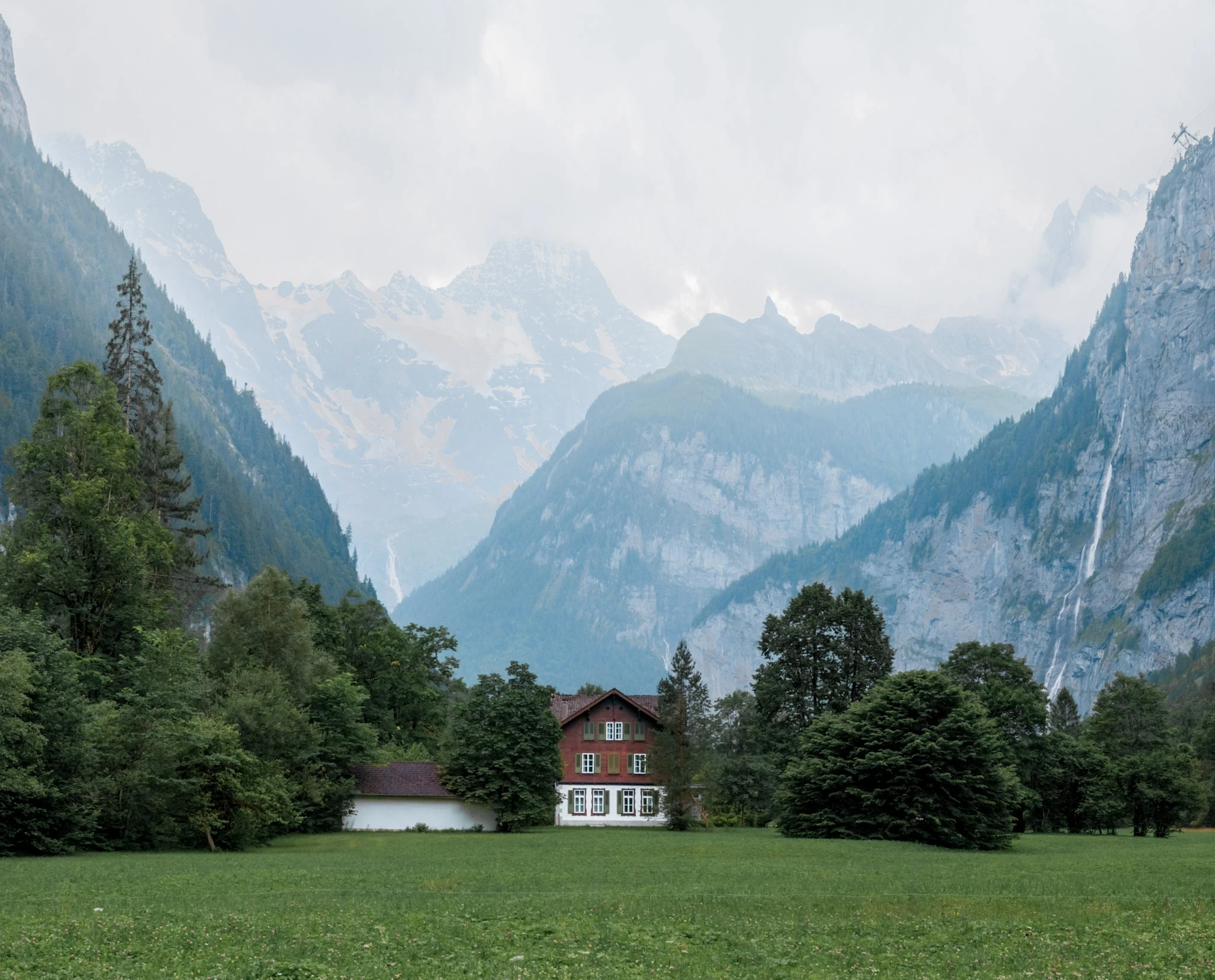 A lone house beneath steep, misty mountains in a green valley.