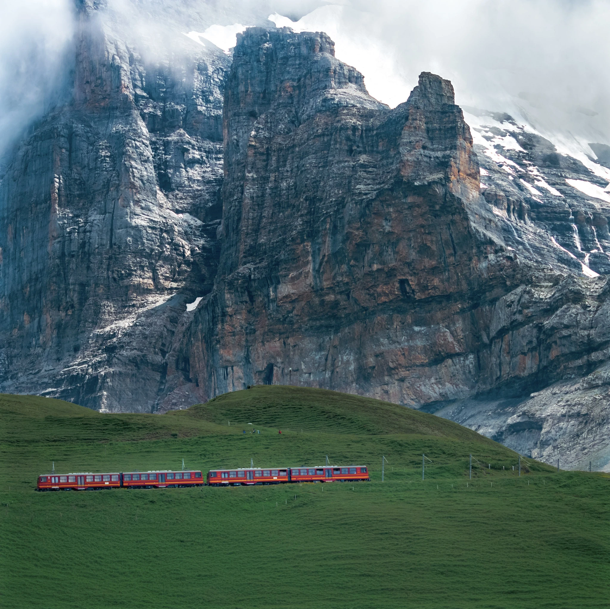 A red mountain train crossing a green meadow below a towering rock face.