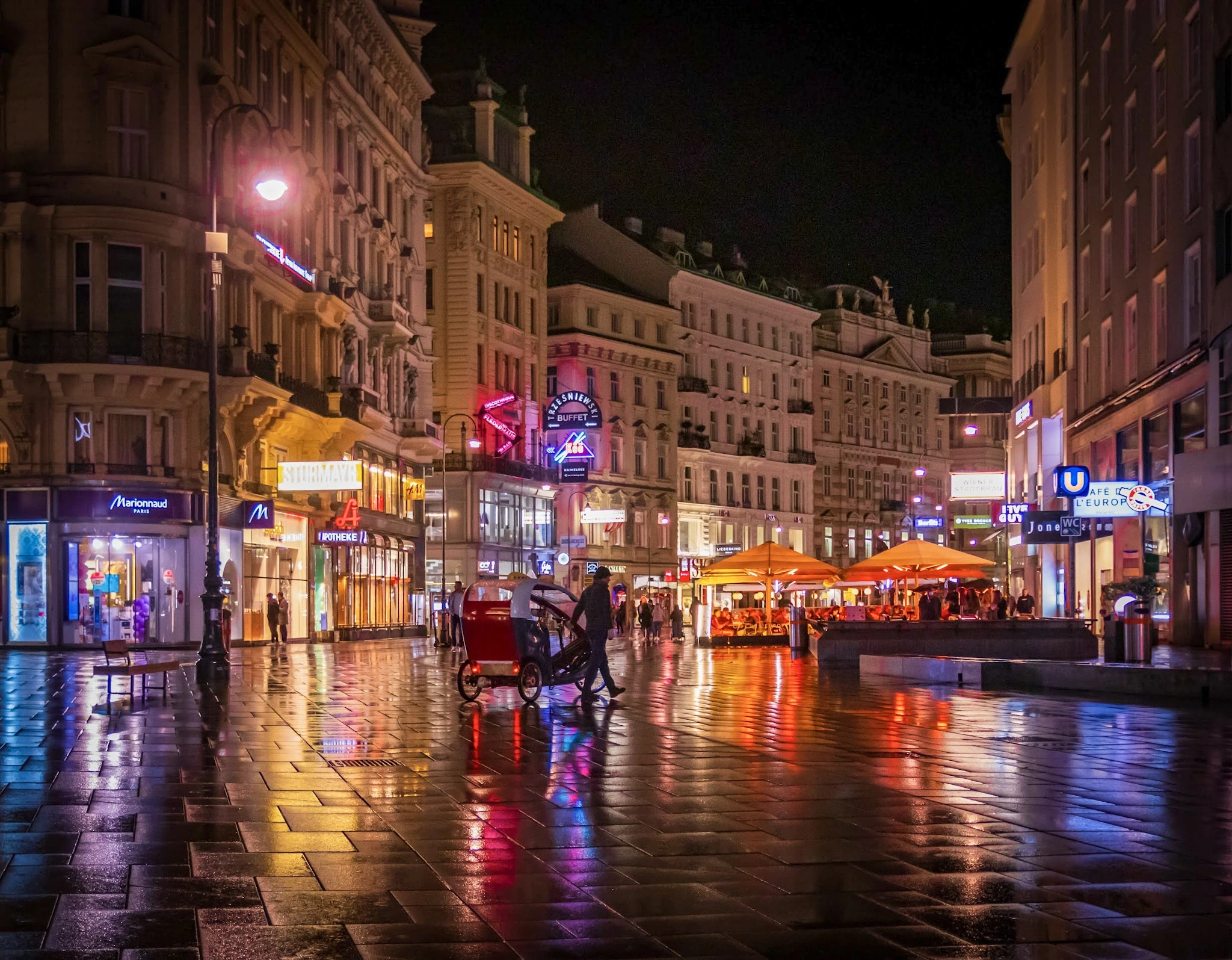 Warm restaurant lights reflected on a rain-soaked city street at night.