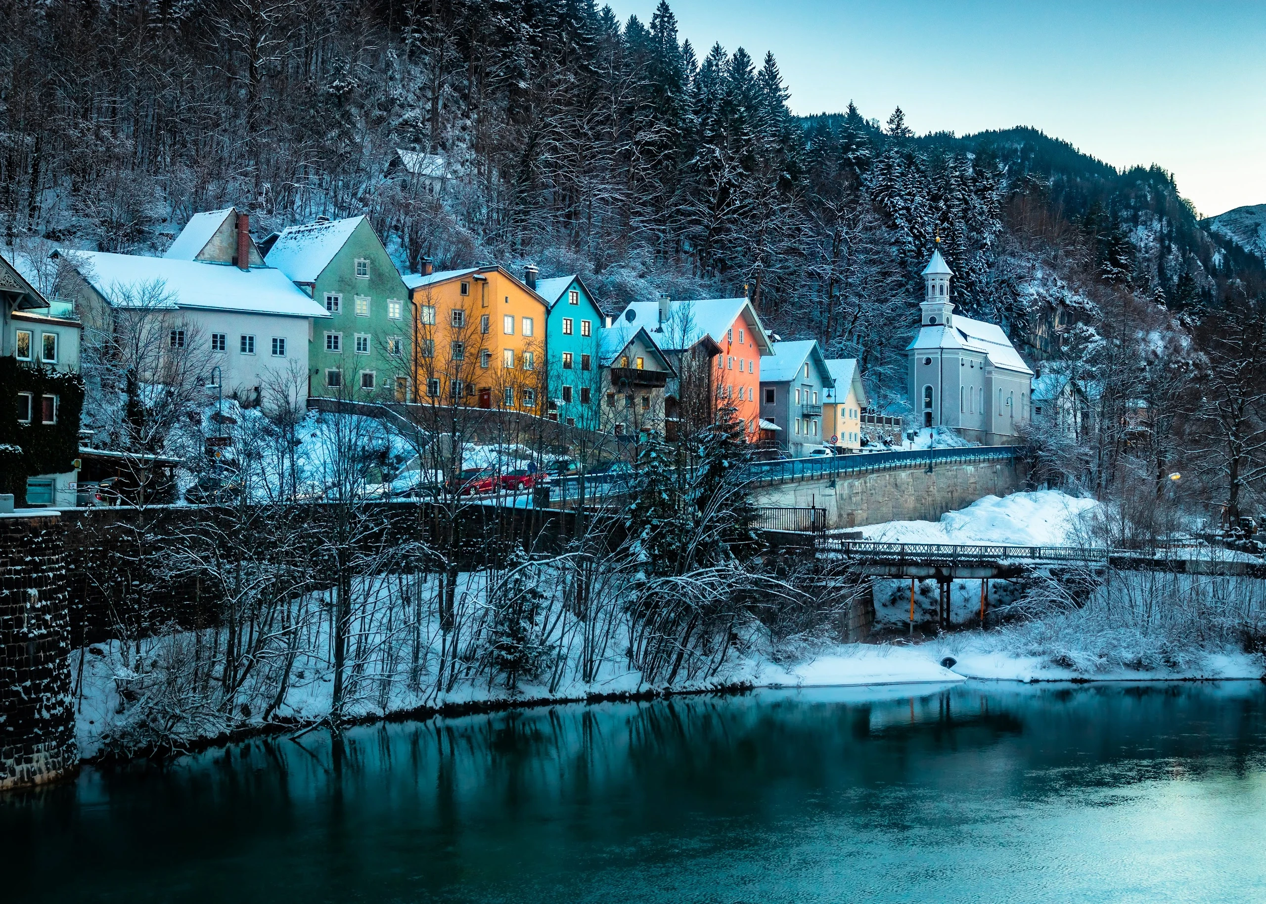 Colourful houses beside a river and snow-dusted forest at blue hour.