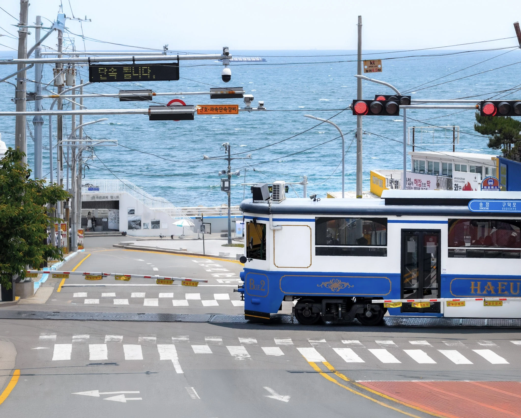 A coastal train at a level crossing with cable cars and blue water beyond.