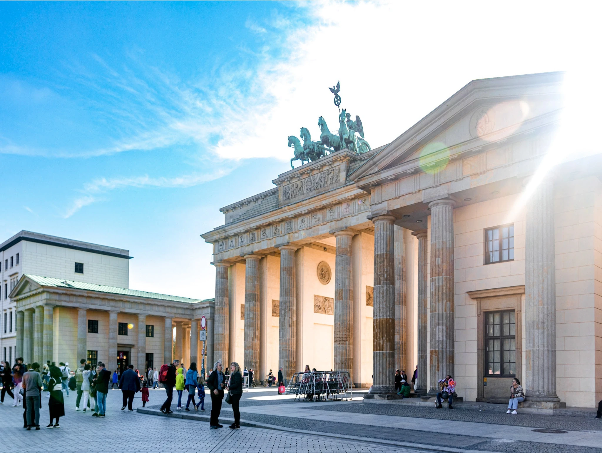 Visitors crossing the square in front of Berlin's Brandenburg Gate in bright sunlight.