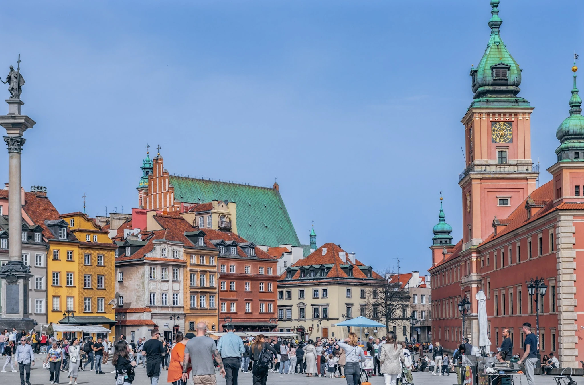 People crossing Warsaw's Castle Square beneath its column and colourful façades.