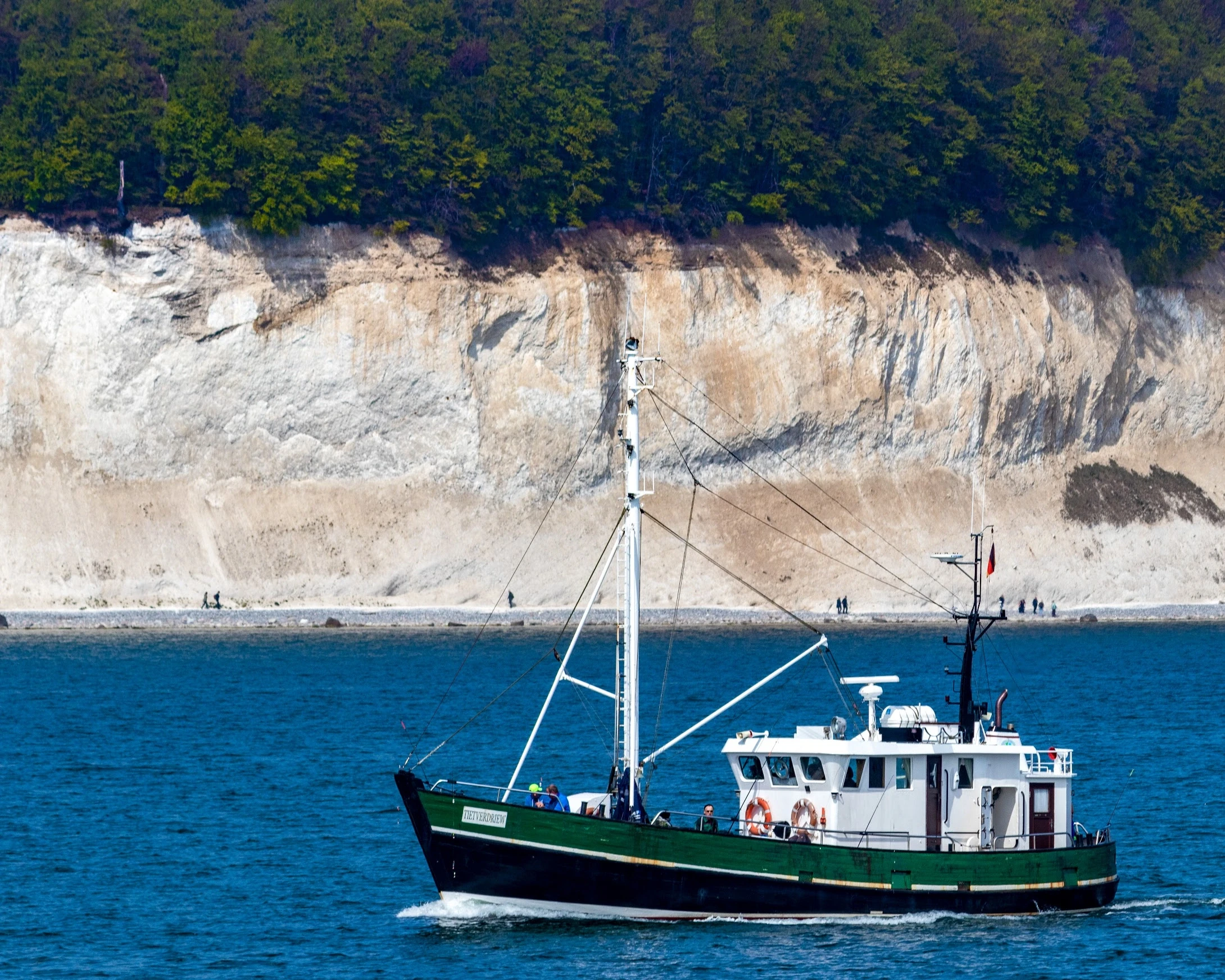 A blue-hulled sailing boat passing white chalk cliffs and green woodland.
