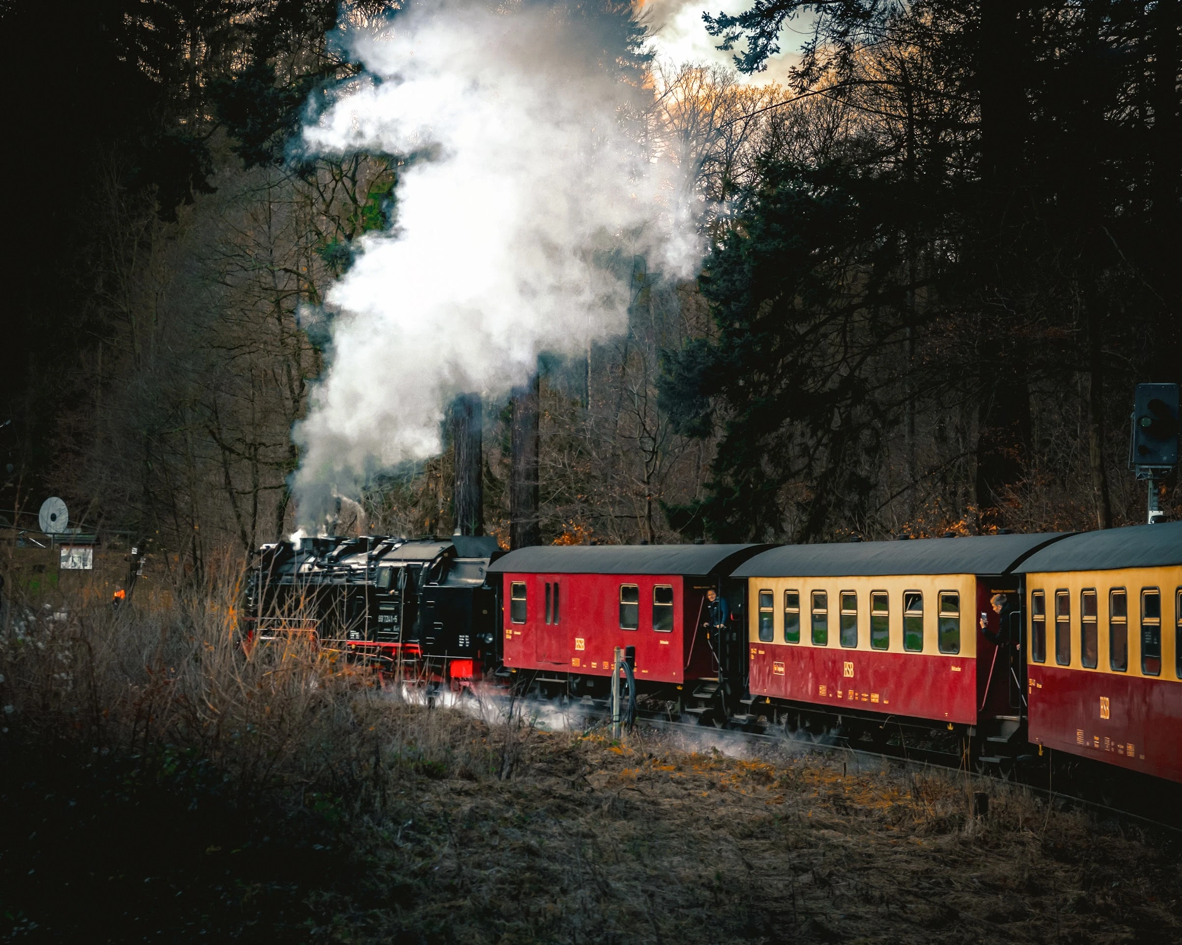 A red steam train sending white smoke through a wooded hillside.