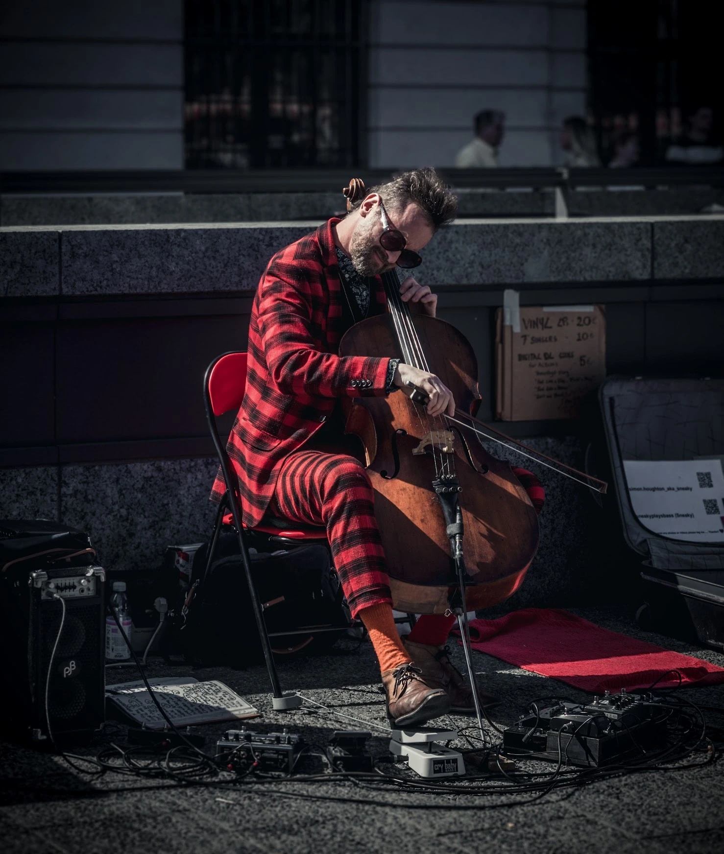A seated guitarist in a red checked shirt playing in a patch of sunlight.
