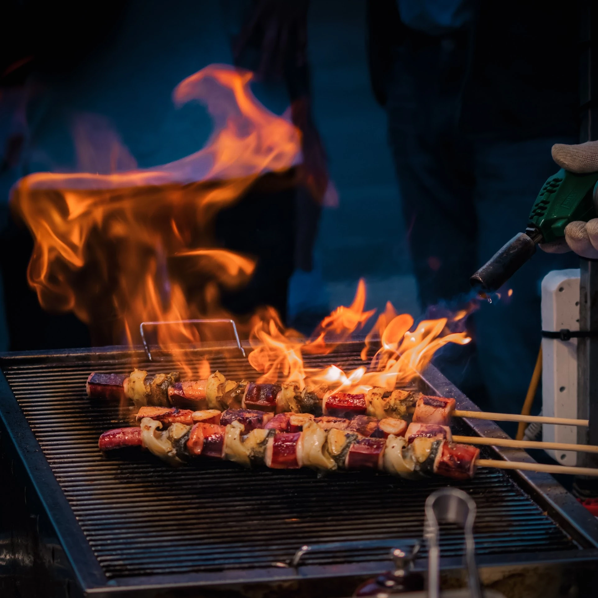 A cook grilling skewers over bright orange flames at a food stall.