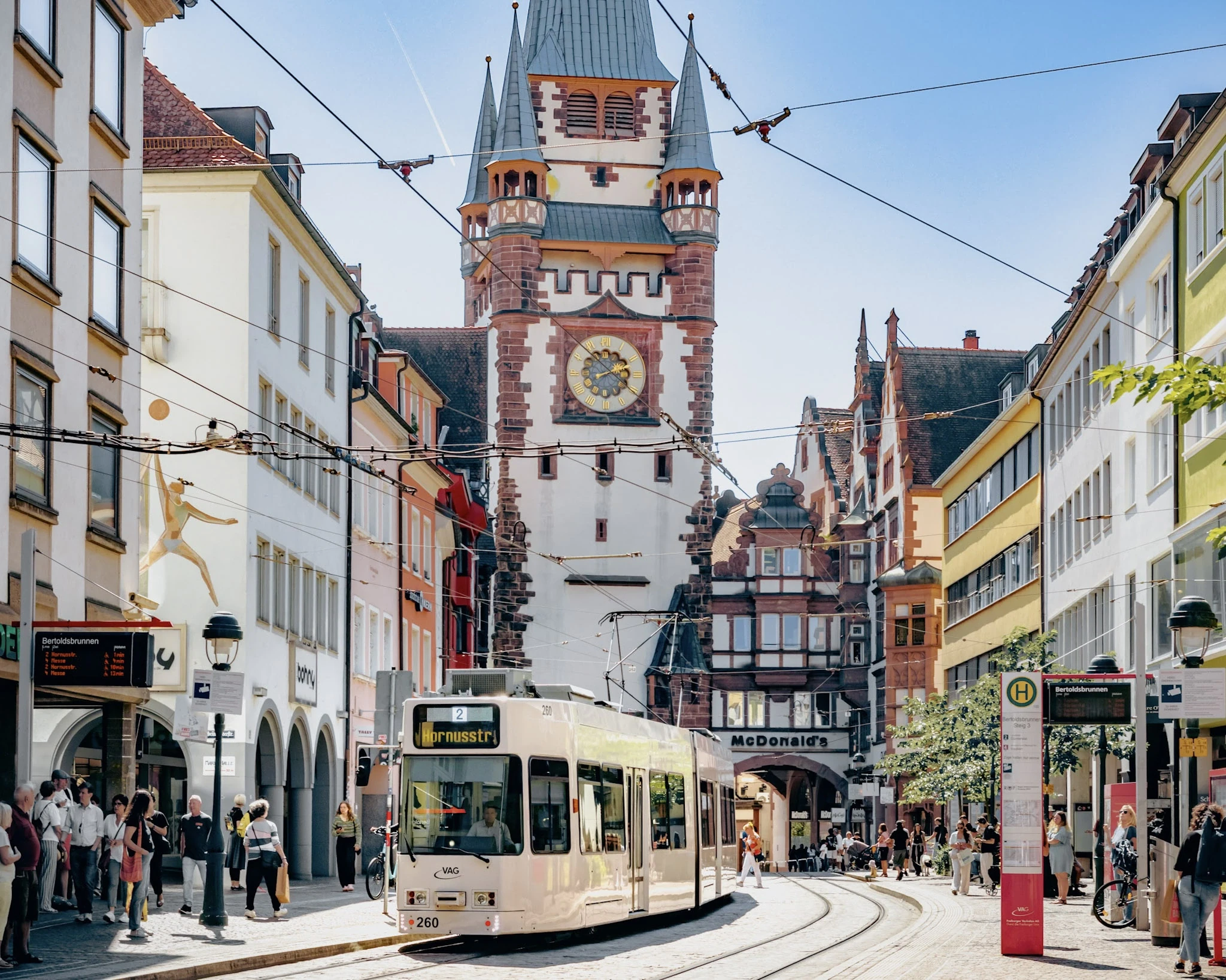 A yellow tram passing a decorated clock tower and colourful old-town façades.