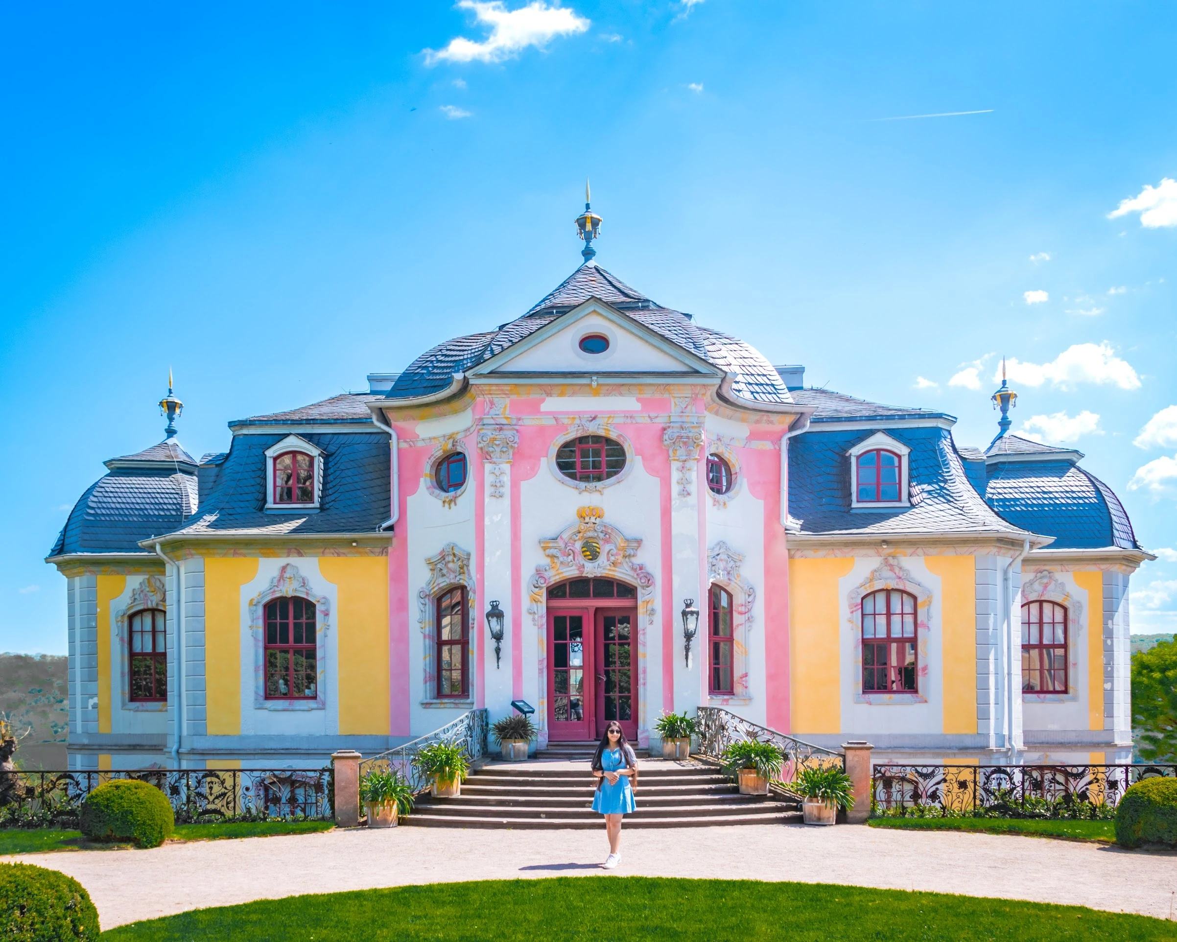 A pink-and-yellow palace façade framed by a formal garden and blue sky.