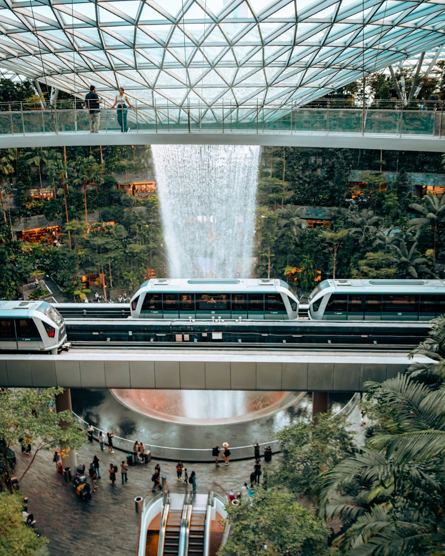 A train passing a waterfall inside a vast glass-roofed indoor garden.