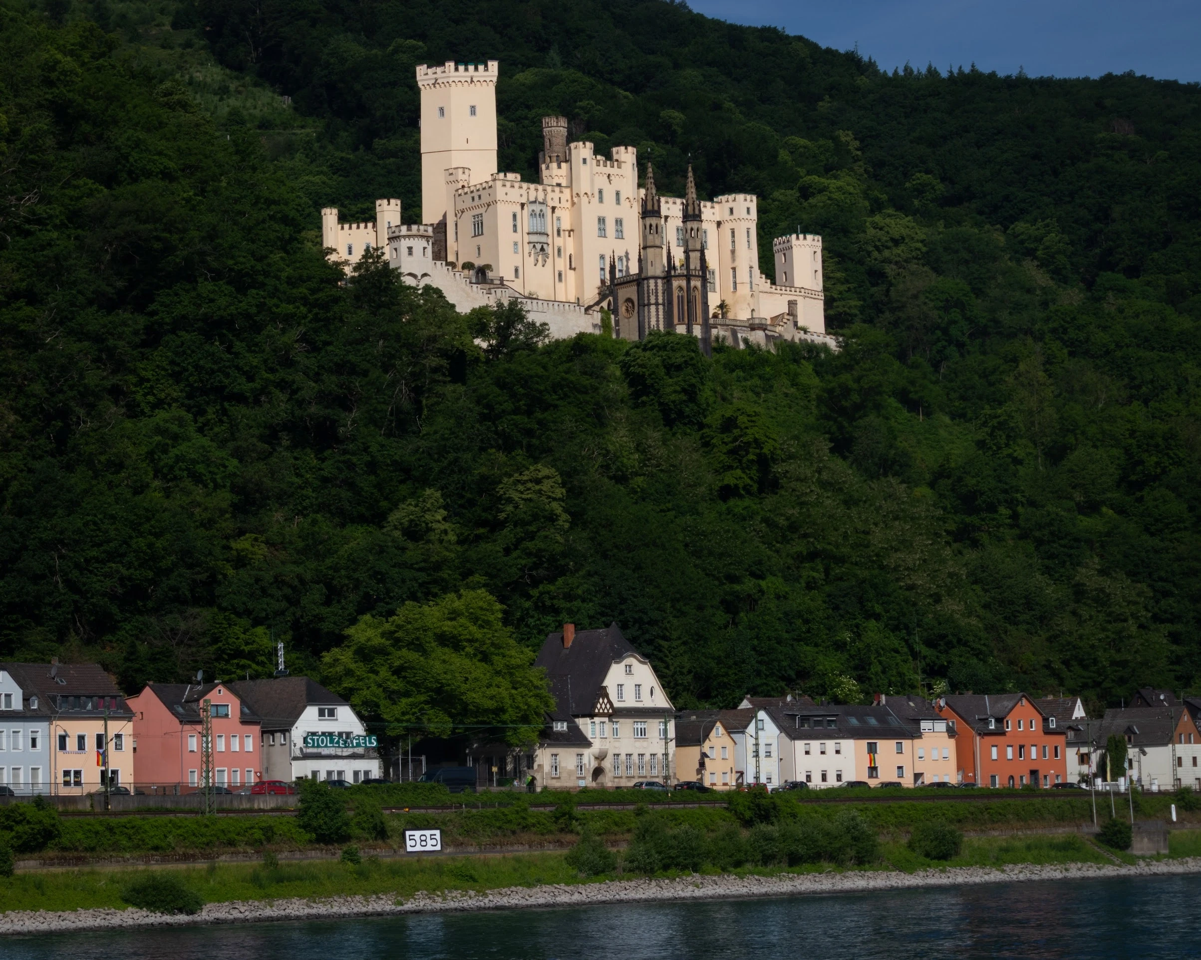 A pale stone castle standing on a forested hill above a riverside village.
