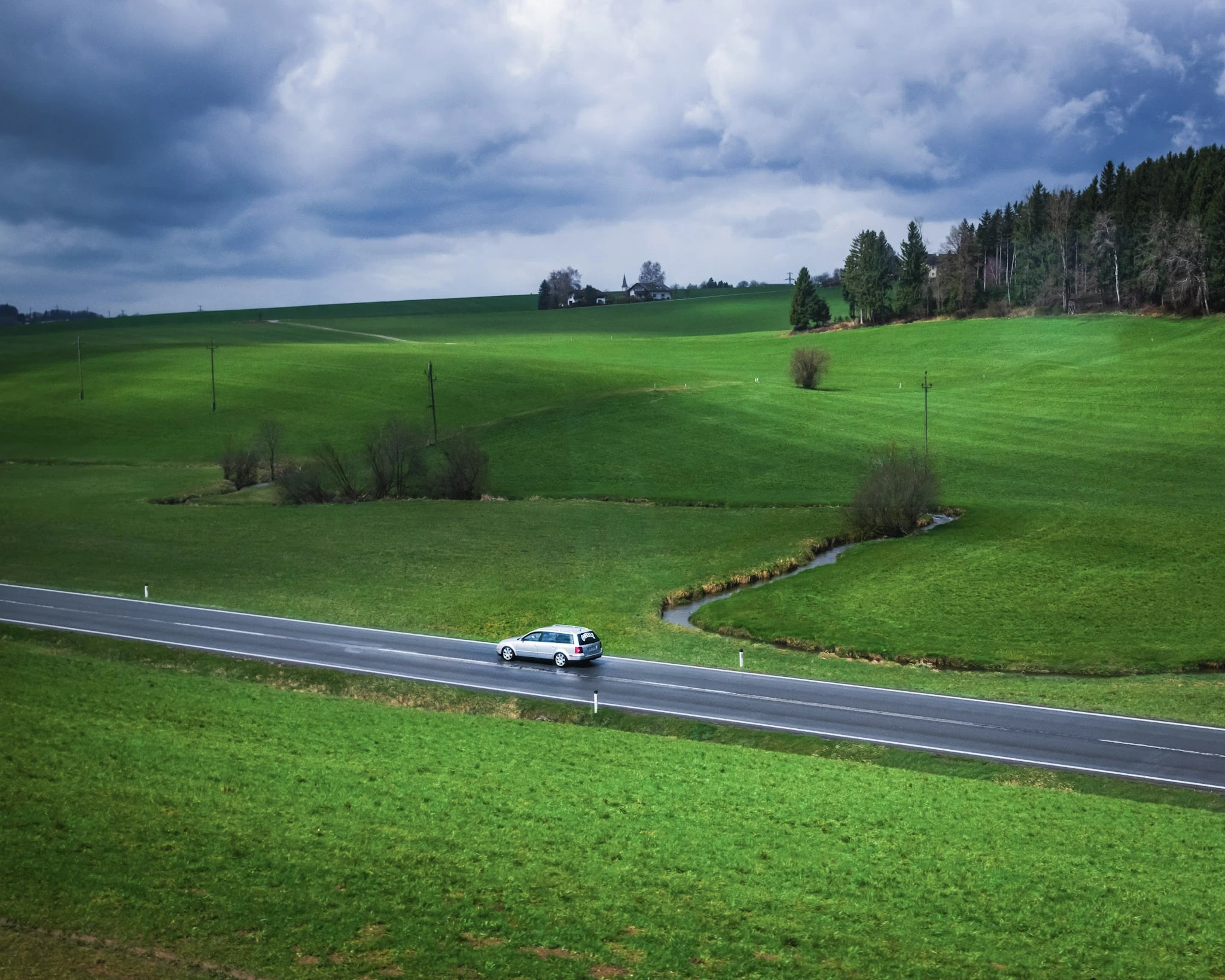 A road winding past bright green fields beneath a dramatic sky.