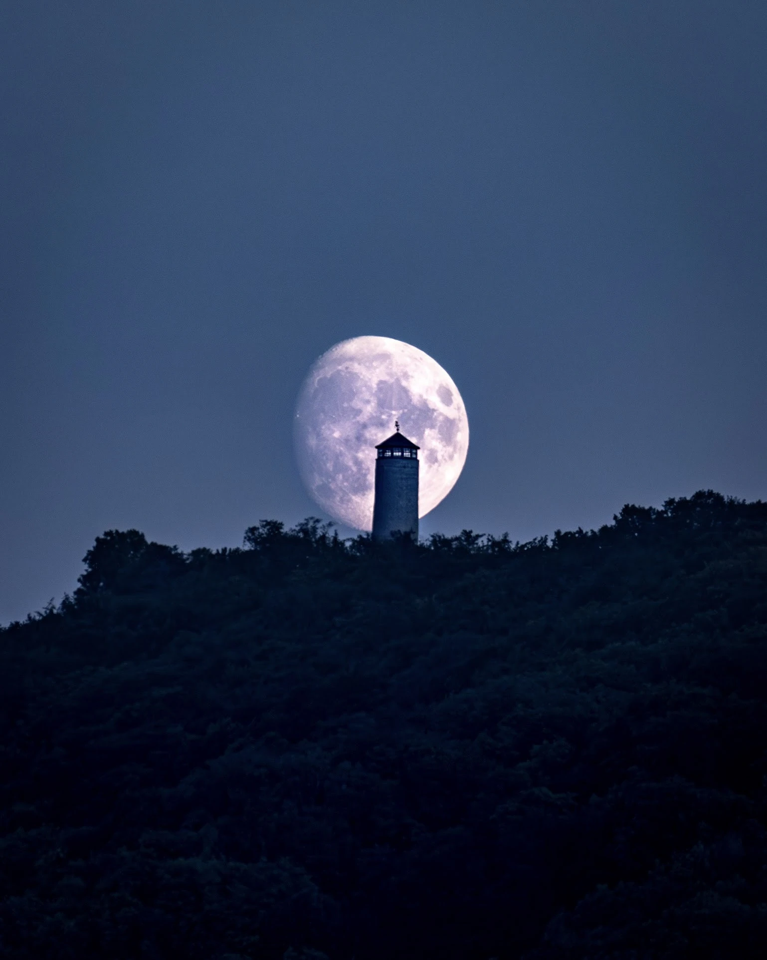 A full moon aligned behind the silhouette of a tower on a dark hillside.