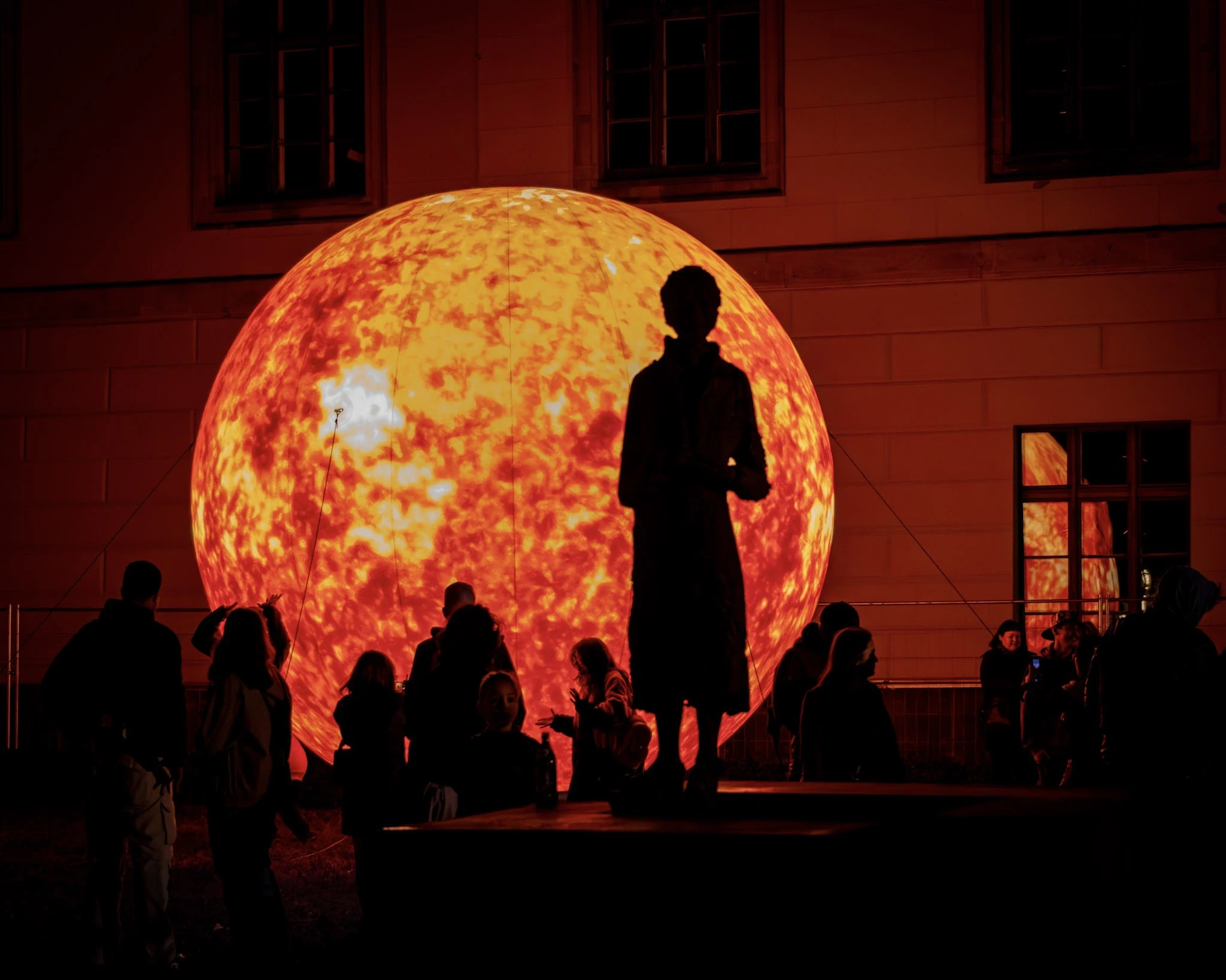 People silhouetted in front of a glowing moon installation beside a city building.