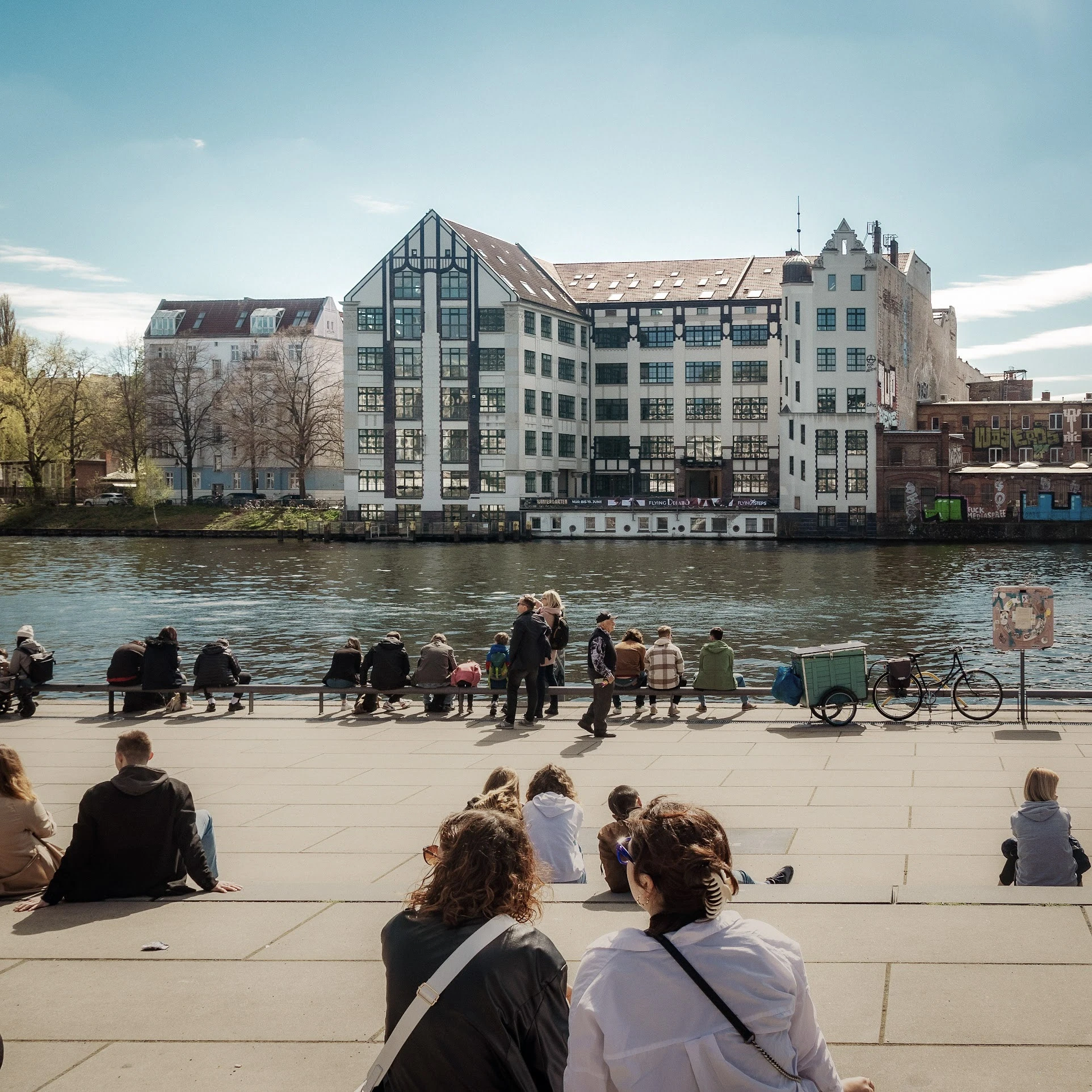 People sitting along a waterfront with modern buildings across the water.